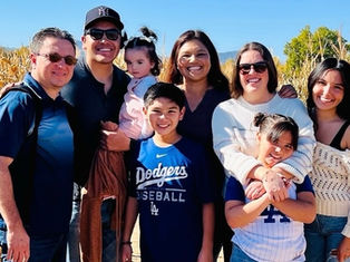 A group of smiling people, including kids, pose outdoors in a cornfield. A boy wears a blue Dodgers T-shirt. Bright, sunny day, clear sky.