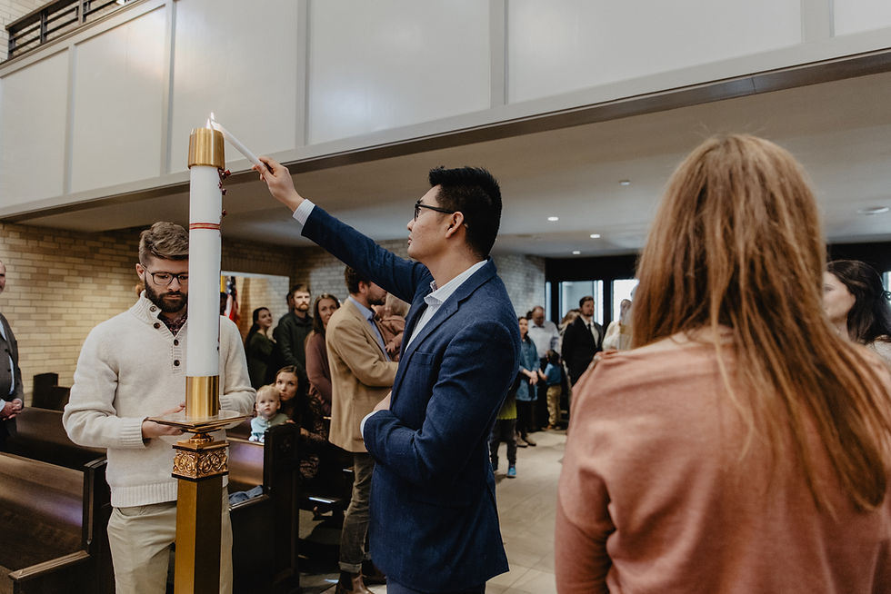 Man in blue blazer lights a tall white candle during a church ceremony while others watch in the pews.