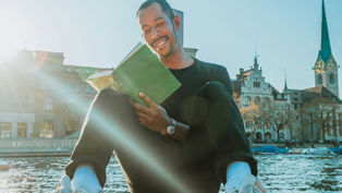 Smiling man in black sits by a lake reading a green book, with sun flare and historic church towers in the background.