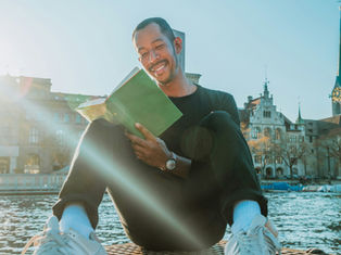 Smiling man in black sits by a lake reading a green book, with sun flare and historic church towers in the background.