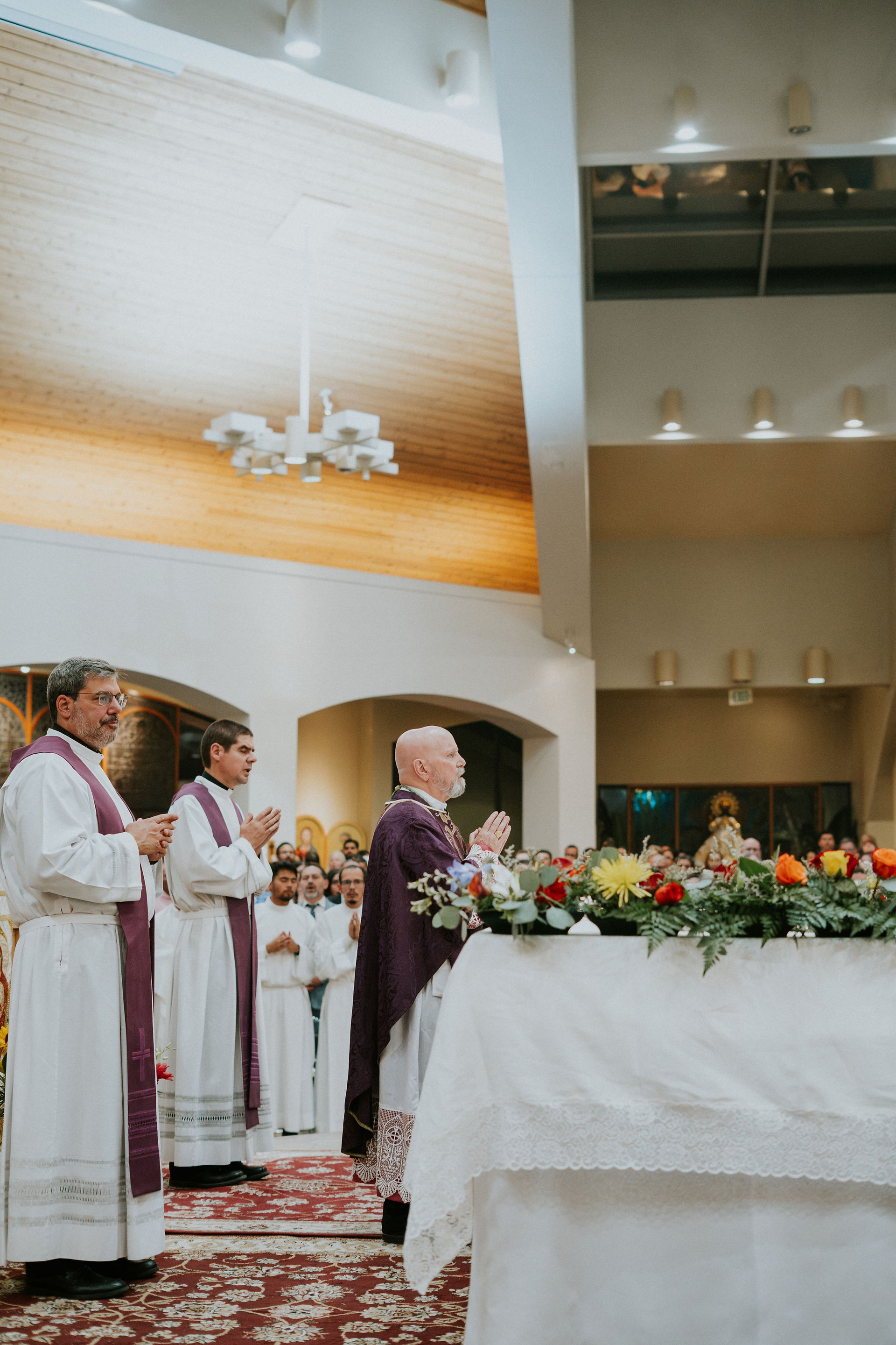Clergy in white and purple robes pray at a flower-adorned altar in a church with a wooden ceiling. Congregation visible in the background.