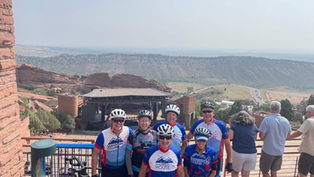 Cyclists in blue and red jerseys pose on a terrace overlooking a scenic rocky amphitheater landscape, with others enjoying the view.