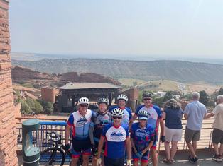Cyclists in blue and red jerseys pose on a terrace overlooking a scenic rocky amphitheater landscape, with others enjoying the view.