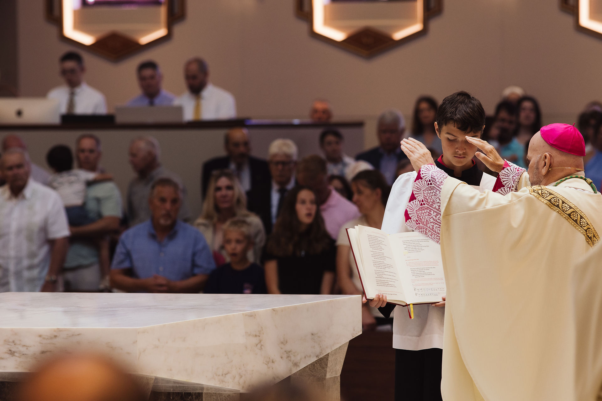 A priest blesses a young acolyte holding a book in a church, with a congregation in the background. The priest wears a pink cap and ornate robe.