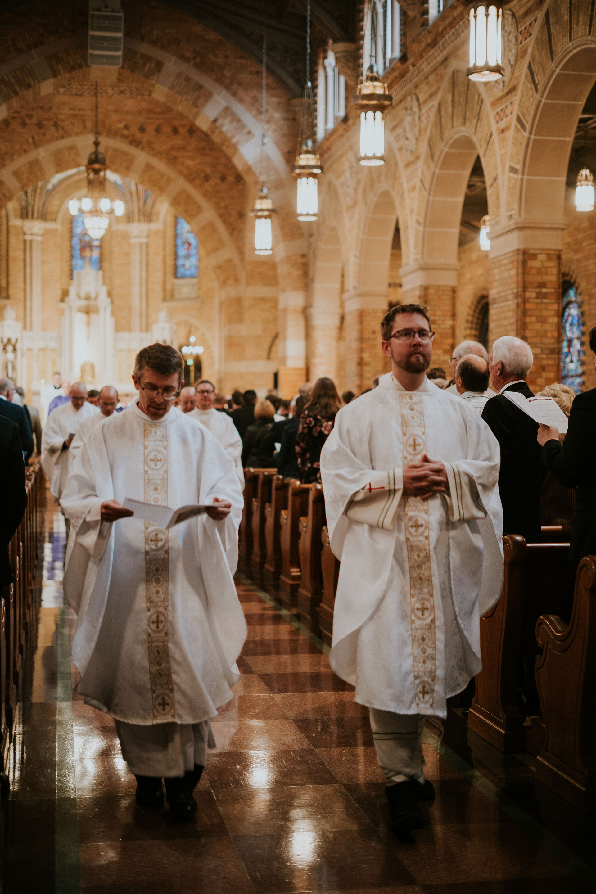 Priests in white robes walk down the aisle of a warmly lit church with high arches and stained glass, holding papers. Ceremony atmosphere.