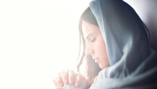A woman in a blue headscarf prays with clasped hands. Her eyes are closed, expressing serenity. Soft lighting creates a peaceful mood.
