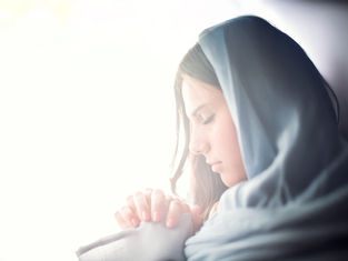 A woman in a blue headscarf prays with clasped hands. Her eyes are closed, expressing serenity. Soft lighting creates a peaceful mood.