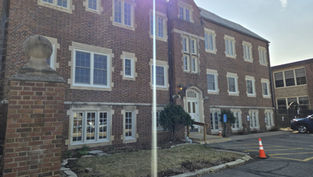Brick building with multiple windows, a white door with "91," and a traffic cone in the parking area. Sunlight casts a glare.