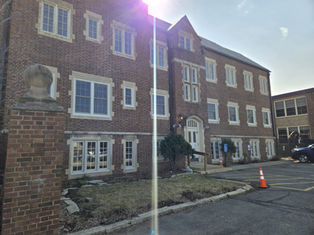 Brick building with multiple windows, a white door with "91," and a traffic cone in the parking area. Sunlight casts a glare.