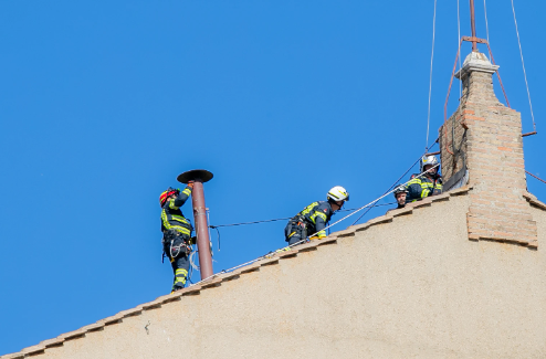 Firefighters in gear work on a rooftop under a clear blue sky, connected by safety ropes. The mood is focused and professional.