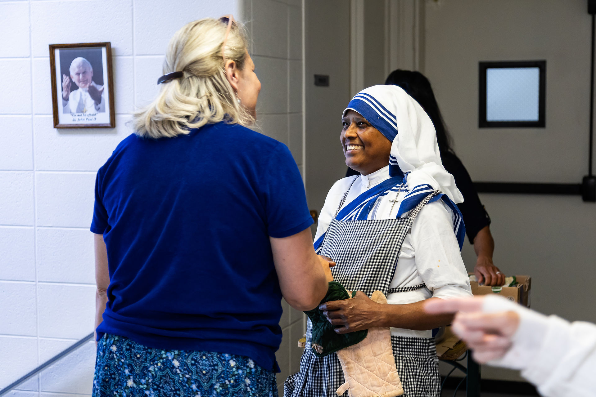 A nun in a blue and white habit smiles warmly at a woman in blue. A framed photo on the wall adds to the cheerful indoor scene.