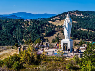Statue of Jesus on a hilltop in a scenic mountainous area. People walk nearby. Green forest and blue sky in the background set a serene mood.