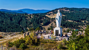 Statue of Jesus on a hilltop in a scenic mountainous area. People walk nearby. Green forest and blue sky in the background set a serene mood.