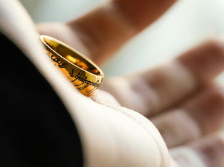 Close-up of a hand holding a golden ring with intricate black script. The background is soft and blurred, creating a contemplative mood.