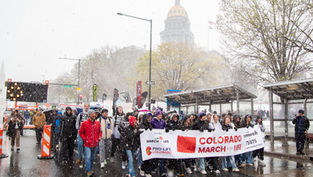 Crowd marches in snowy weather holding "Colorado March for Life" banner. Capitol building in background. People dressed warmly, mood is determined.