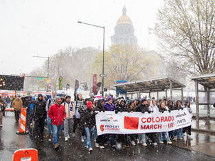 Crowd marches in snowy weather holding "Colorado March for Life" banner. Capitol building in background. People dressed warmly, mood is determined.