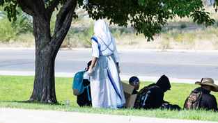 Person in a white and blue religious habit stands near seated people under a tree. Backpacks and boxes are visible on a grassy area.