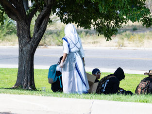 Person in a white and blue religious habit stands near seated people under a tree. Backpacks and boxes are visible on a grassy area.