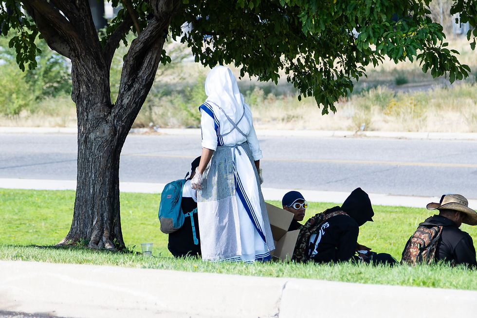 Person in a white and blue religious habit stands near seated people under a tree. Backpacks and boxes are visible on a grassy area.