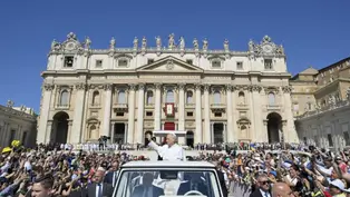 A white-clad figure waves from a vehicle in front of a grand cathedral, greeted by a crowd. Clear blue sky and vibrant atmosphere.