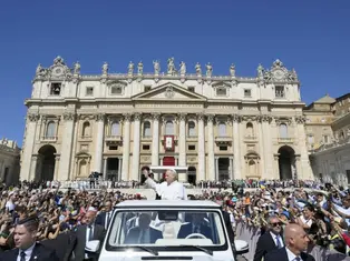 A white-clad figure waves from a vehicle in front of a grand cathedral, greeted by a crowd. Clear blue sky and vibrant atmosphere.