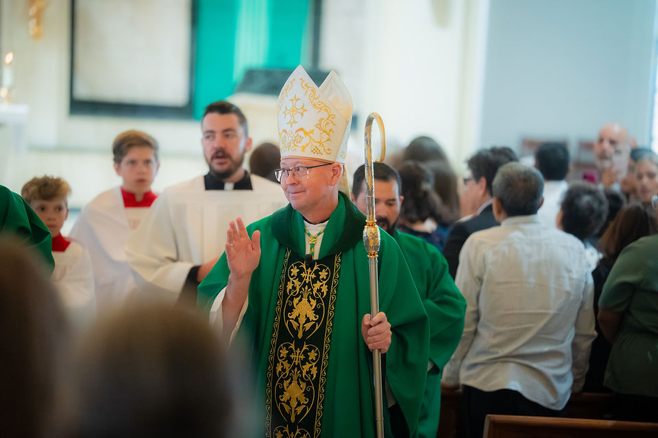 Bishop in green vestments and gold mitre waves while holding a crosier during a church procession, with altar servers and crowd behind.