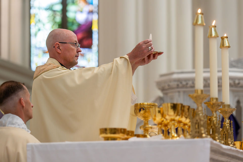 Bishop holding up a communion wafer during Mass in a church. Candles and golden chalices on the altar, stained glass window in background.