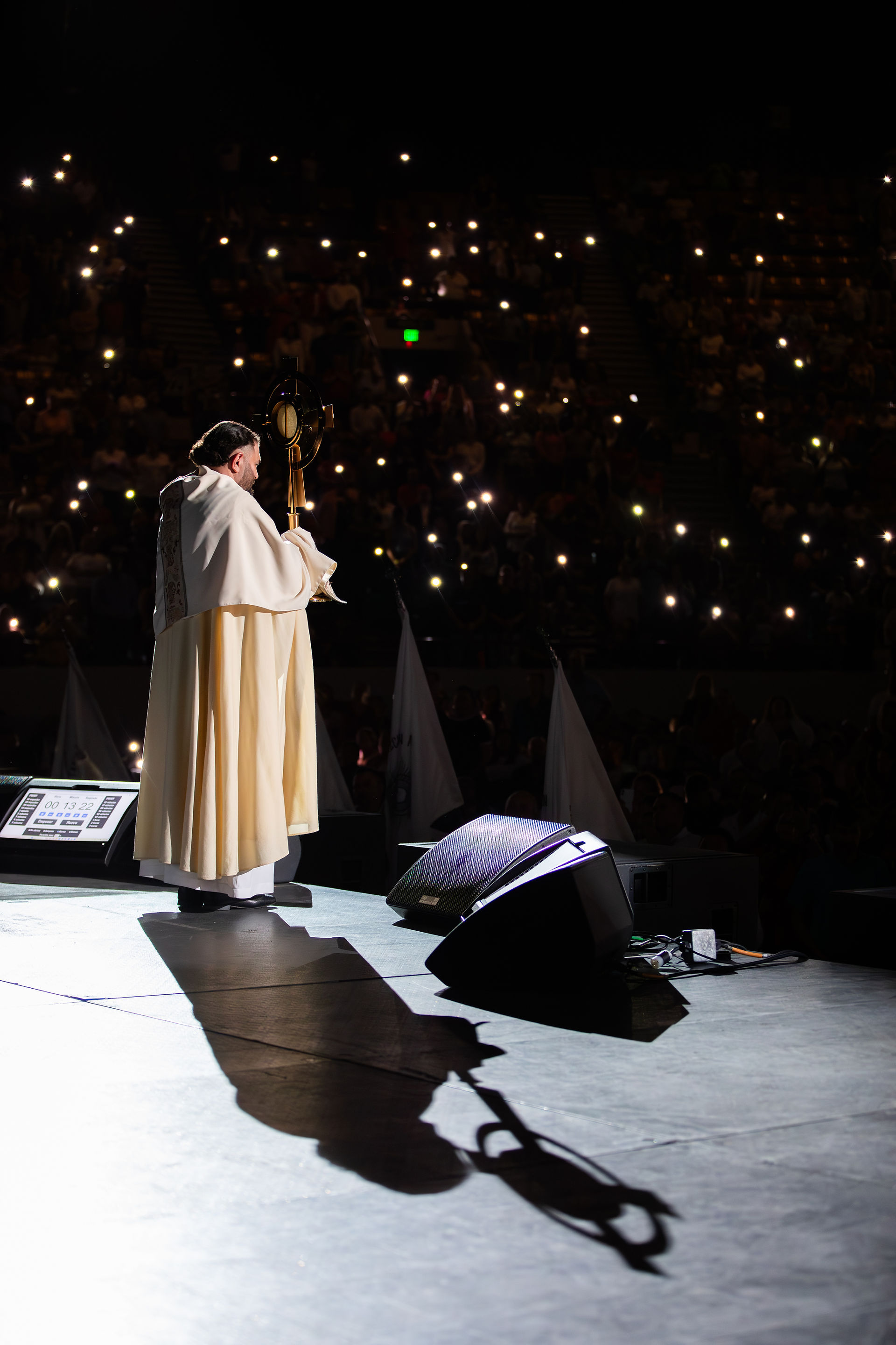 Priest in light robe holds up a religious object on stage. Audience with phone lights in dark background creates a solemn atmosphere.
