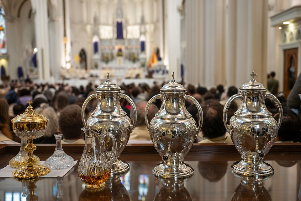 Silver vessels and a glass pitcher in a cathedral with a blurred congregation and ornate altar in the background, creating a solemn atmosphere.