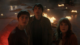 Three people look startled in a dim, fiery setting with scattered fires. A vehicle's headlights are visible in the smoky background.