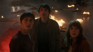 Three people look startled in a dim, fiery setting with scattered fires. A vehicle's headlights are visible in the smoky background.