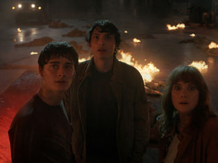 Three people look startled in a dim, fiery setting with scattered fires. A vehicle's headlights are visible in the smoky background.