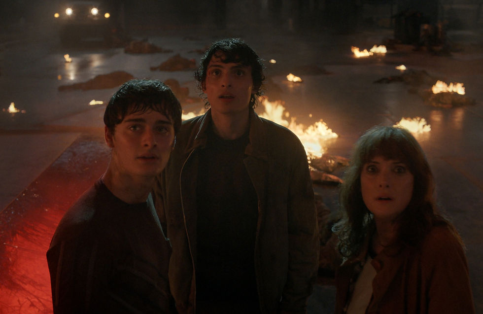Three people look startled in a dim, fiery setting with scattered fires. A vehicle's headlights are visible in the smoky background.