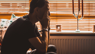 A person sits in profile, hands on face, silhouetted against orange blinds. Mood appears contemplative. Candles and patterned pillows visible.