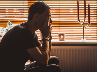 A person sits in profile, hands on face, silhouetted against orange blinds. Mood appears contemplative. Candles and patterned pillows visible.