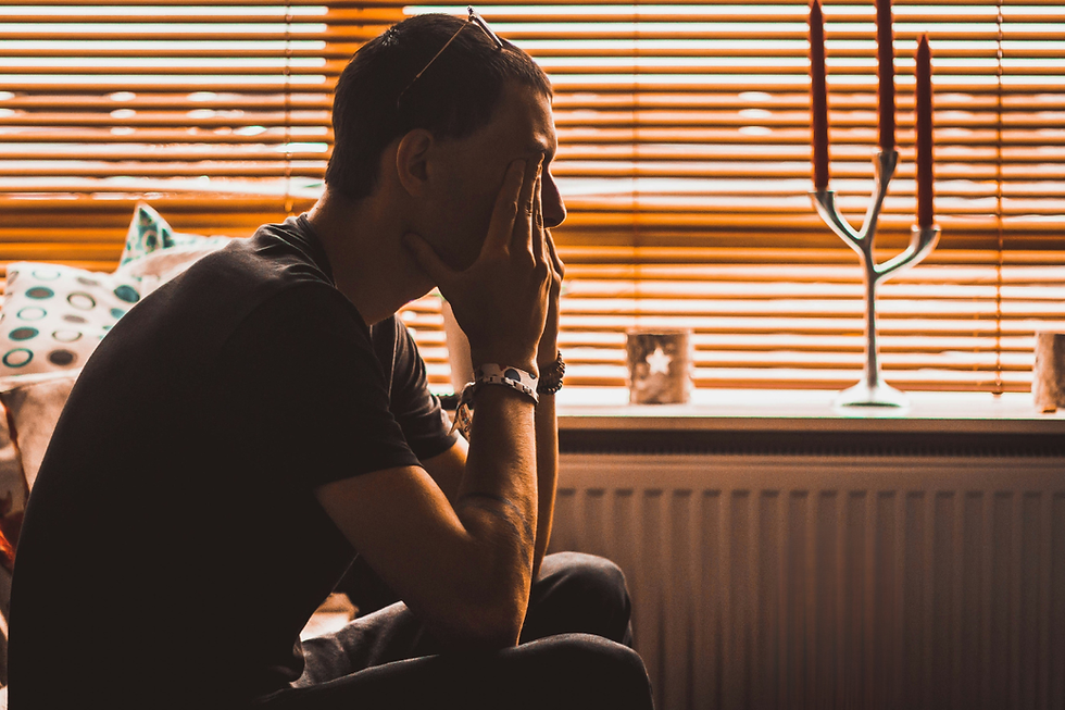 A person sits in profile, hands on face, silhouetted against orange blinds. Mood appears contemplative. Candles and patterned pillows visible.