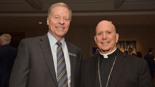 Two men in suits smile at a formal event. The background has patterned artwork, and a sign with text is partially visible. Atmosphere is friendly.