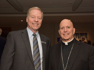 Two men in suits smile at a formal event. The background has patterned artwork, and a sign with text is partially visible. Atmosphere is friendly.