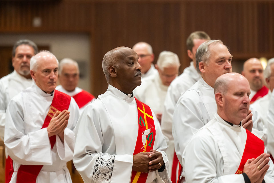 Clergymen in white robes with red stoles pray in a church. The setting is serene, and their expressions are solemn and focused.