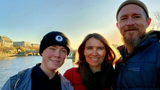 Three people smiling near a river with buildings and a bridge in the background at sunset. Two wear beanies, one wears a blue jacket.