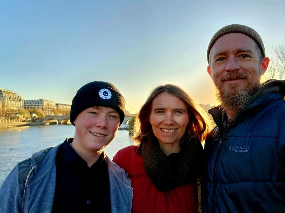 Three people smiling near a river with buildings and a bridge in the background at sunset. Two wear beanies, one wears a blue jacket.