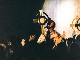 Catholic priest holds a golden monstrance before raised hands in a dim, glowing worship service.