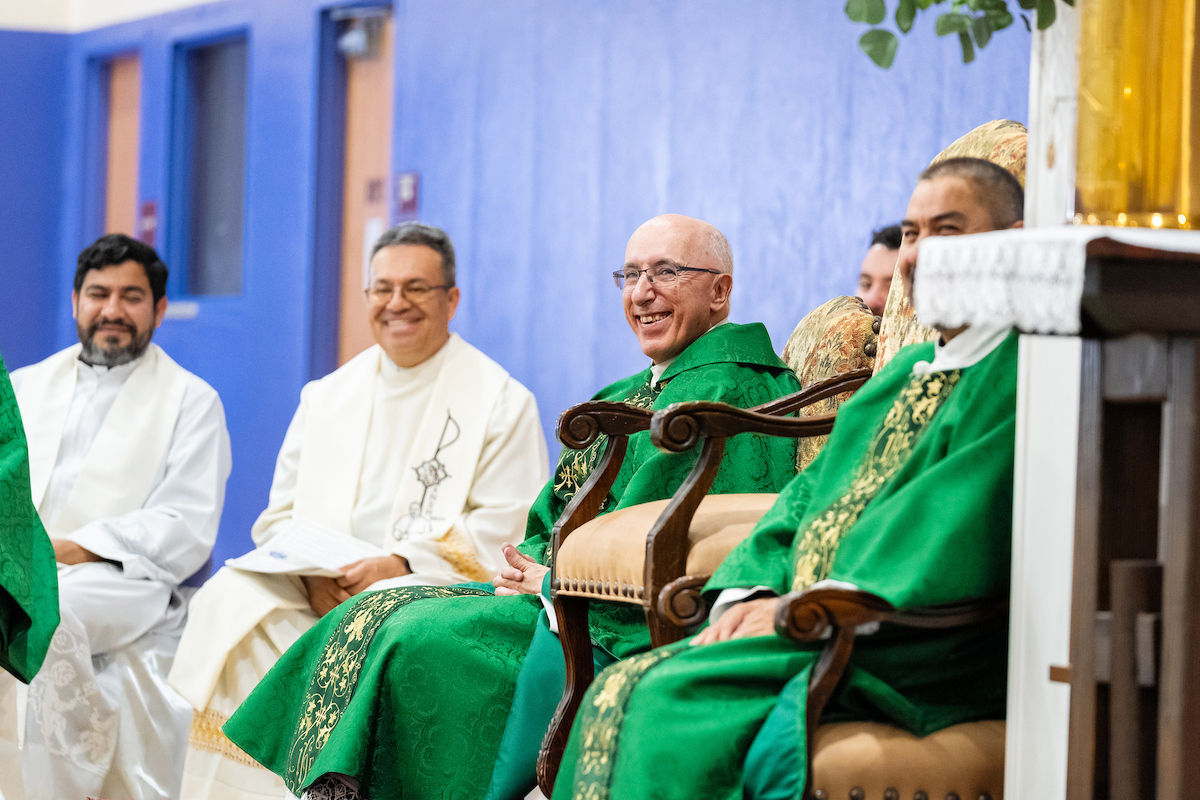 Smiling priests in green and white vestments sit in a church hall against a blue wall during a religious ceremony.
