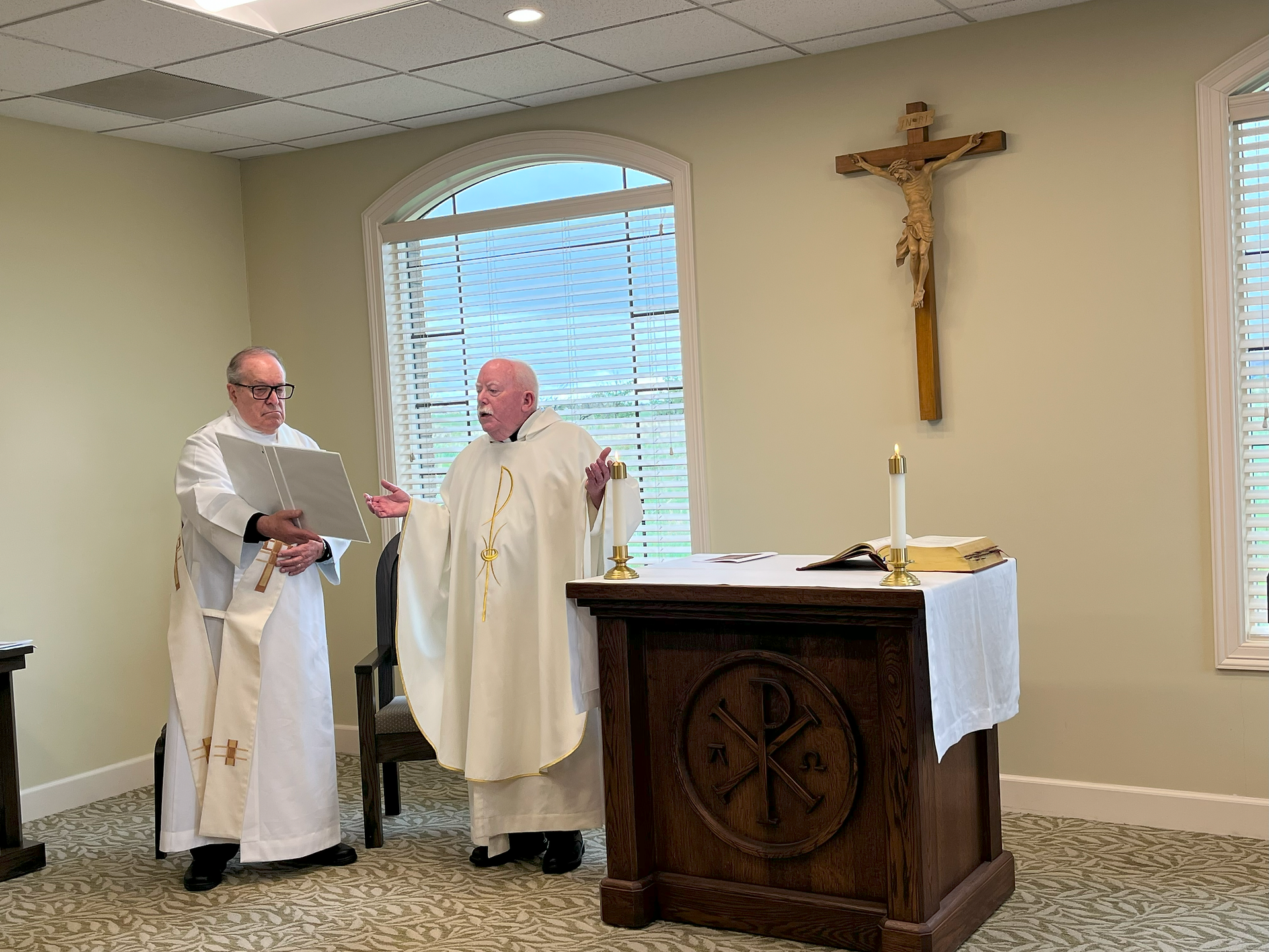 Two clergy in white robes stand by an altar. One reads from a book, the other gestures. A crucifix hangs on the wall. Calm setting.