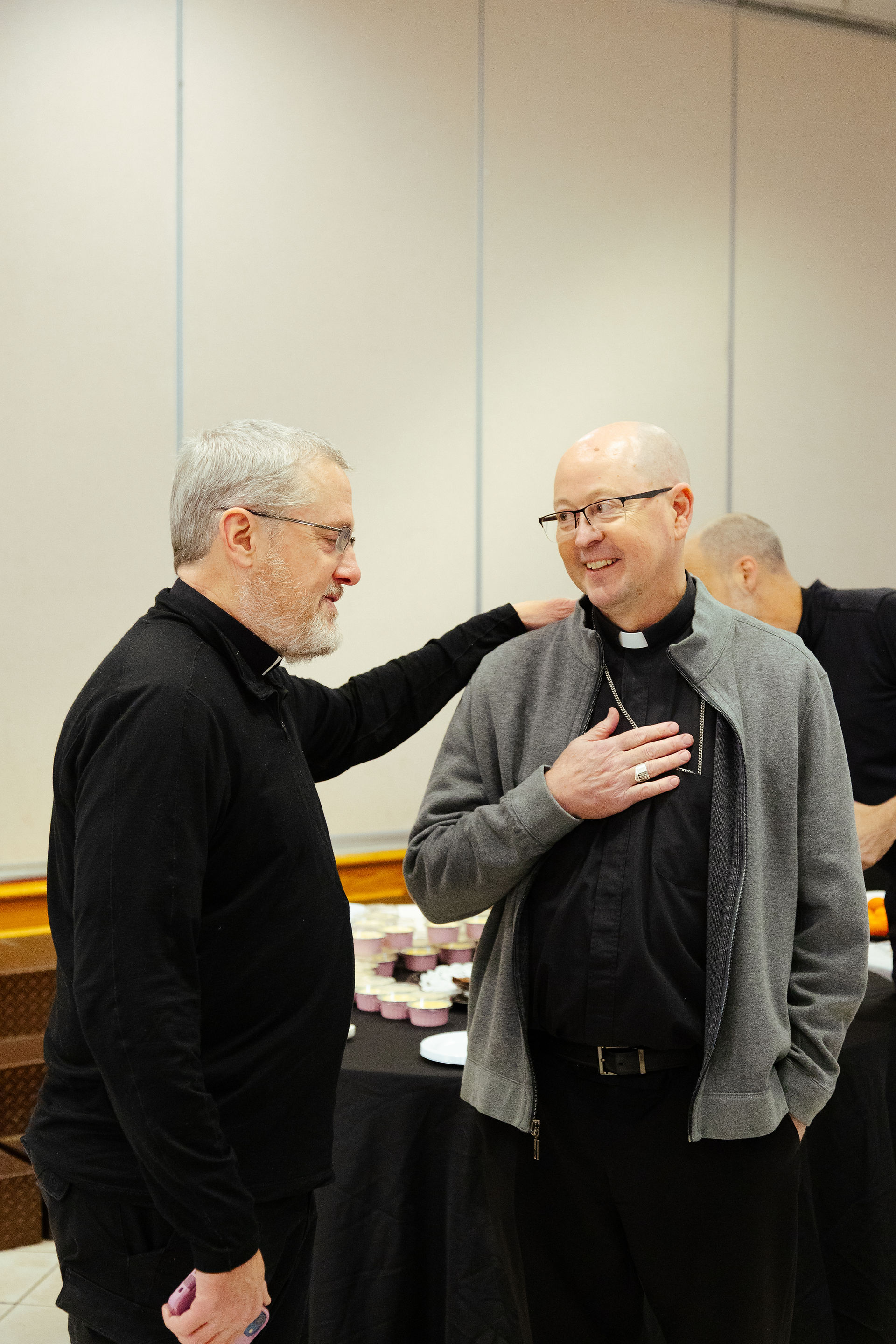 Two men in clerical attire converse warmly, one with a hand on the other's shoulder. Background shows a table with cups and plates.