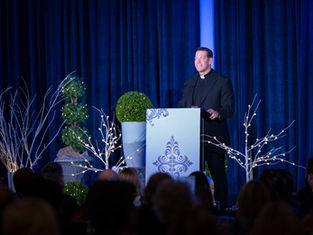 Man in black clerical attire speaks at a lectern with decorative designs. Blue curtains and lit branches set a formal, serene mood.