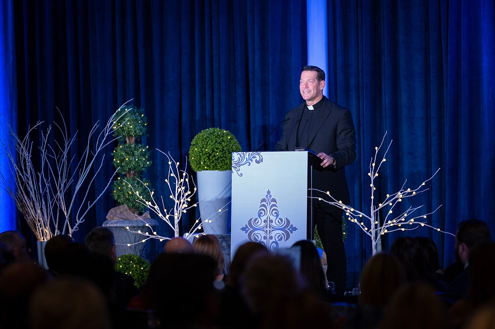 Man in black clerical attire speaks at a lectern with decorative designs. Blue curtains and lit branches set a formal, serene mood.
