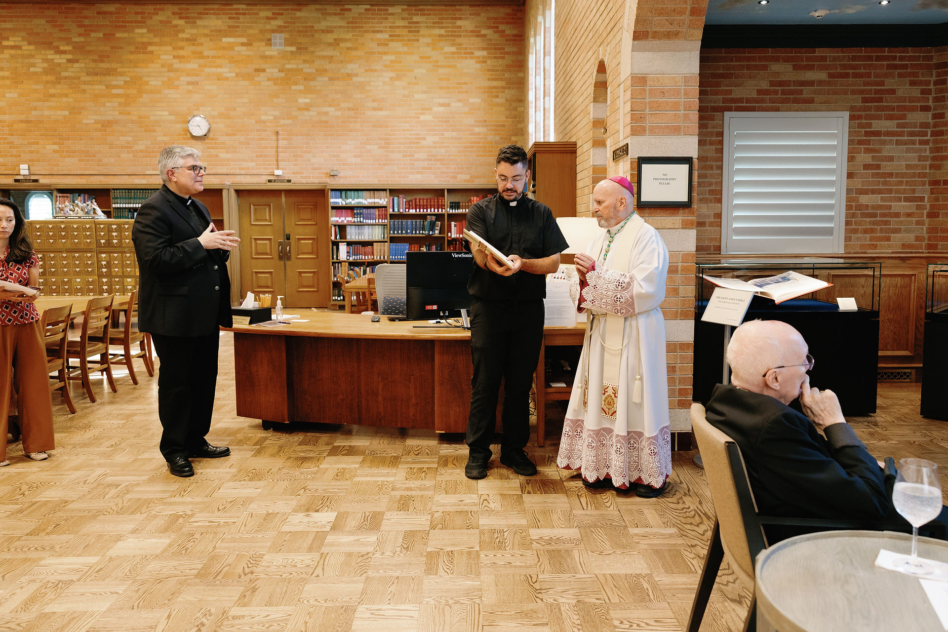 Priest shows a book to a bishop in a library with brick walls. Two others observe, one seated. Shelves and wooden furniture in background.