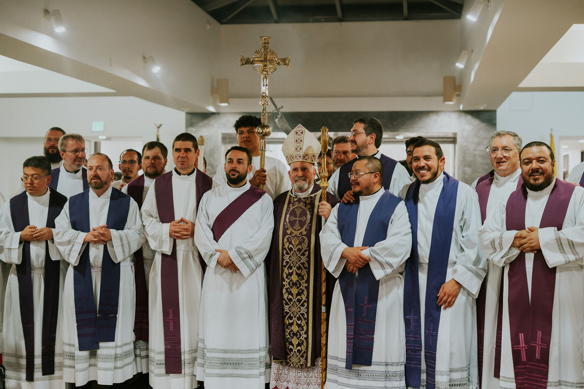 A group of clergy in purple and white robes stands together in a church. The bishop in the center holds a gold cross, smiling.