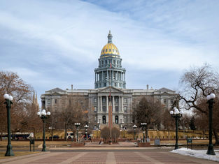 Gray domed capitol building with a golden top against a cloudy sky. Trees and streetlights line the walkway. Sparse visitors in the foreground.